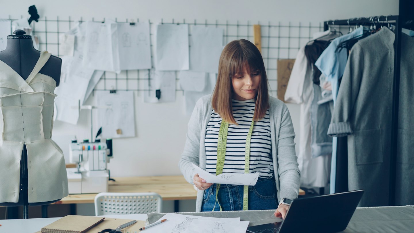 Fashion designer reviews sketches at a desk with a laptop.